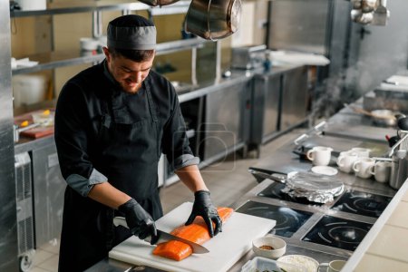 Téléchargez les photos : Cuisine professionnelle dans le restaurant de l'hôtel chef coupe une grande carcasse de saumon frais avec un couteau - en image libre de droit