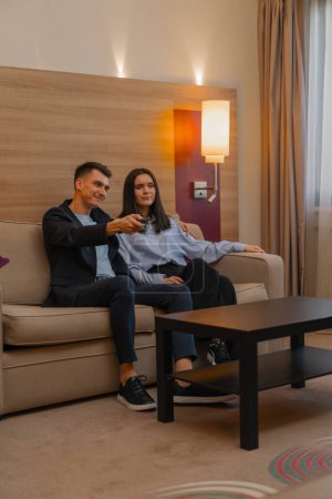Téléchargez les photos : Jeune couple chambre d'hôtel salon regarder la télévision ensemble L'homme prend la télécommande et change de chaînes - en image libre de droit
