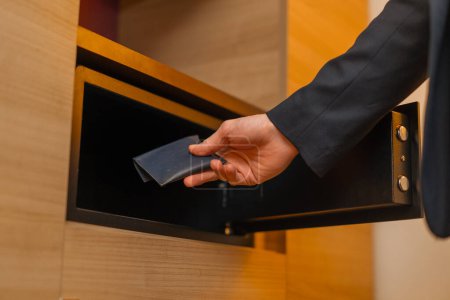 Téléchargez les photos : Gros plan de la main d'un homme mettant un portefeuille avec de l'argent dans un coffre-fort dans la chambre d'hôtel stockage des valeurs - en image libre de droit