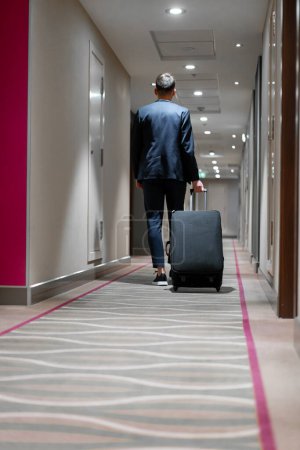 Téléchargez les photos : Un homme avec une valise marche dans le couloir de l'hôtel une vue arrière d'un homme d'affaires en voyage - en image libre de droit