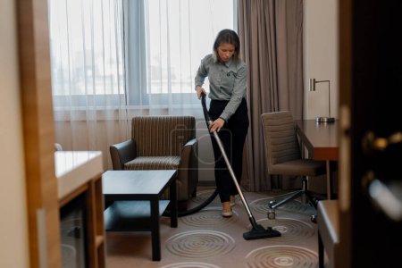 Foto de Una sirvienta diligente en uniforme cuidadosamente aspira la alfombra mientras limpia la habitación del hotel - Imagen libre de derechos