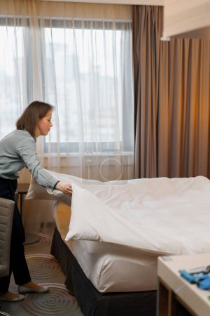 Téléchargez les photos : Une femme de ménage en uniforme fait un lit préparant une chambre d'hôtel de luxe pour les clients nettoyage et Voyage concept - en image libre de droit