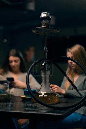 Foto de Dos amigas jóvenes están sentadas relajadas en el bar de narguile fumando narguile y divirtiéndose charlando concepto de recreación - Imagen libre de derechos