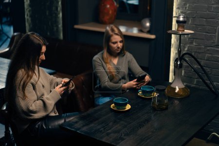 Foto de Dos novias jóvenes están sentadas relajadas en el bar de narguile con teléfonos en sus manos concepto de descanso - Imagen libre de derechos