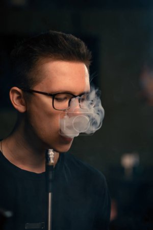 Foto de Un hombre de narguile con gafas fuma una pipa de narguile tradicional Un hombre exhala humo en la cafetería de narguile o en el bar salón de cerca - Imagen libre de derechos