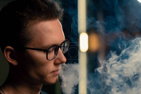 Foto de Un hombre de narguile con gafas fuma una pipa de narguile tradicional Un hombre exhala humo en la cafetería de narguile o en el bar salón de cerca - Imagen libre de derechos