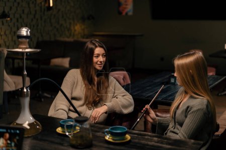 Foto de Dos amigas jóvenes están sentadas relajadas en el bar de narguile fumando narguile y divirtiéndose charlando concepto de recreación - Imagen libre de derechos