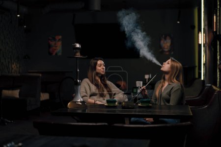 Photo for Two young female friends are sitting relaxed in hookah bar smoking hookah and having fun chatting recreation concept - Royalty Free Image