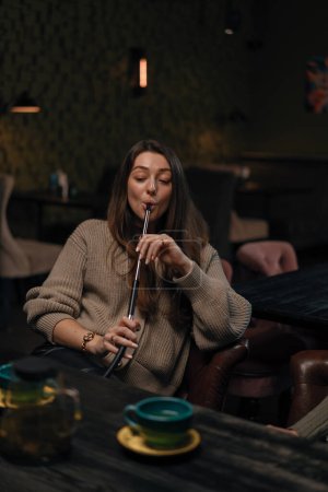 Photo for Beautiful young woman inhaling hookah girl smoking hookah sitting in lounge bar concept relaxation and smoking - Royalty Free Image