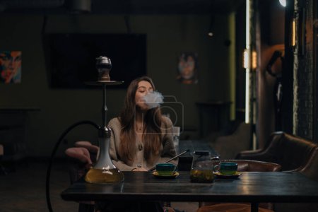 Foto de Hermosa mujer joven inhalando cachimba chica fumando cachimba sentado en salón bar concepto relajación y fumar - Imagen libre de derechos