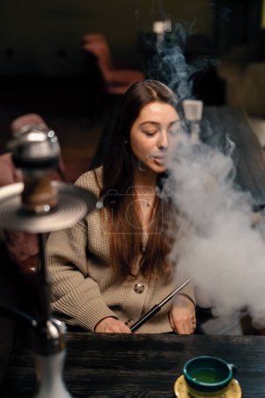 Téléchargez les photos : Belle jeune femme inhalant narguilé fille fumeur narguilé assis dans le concept de bar salon détente et fumer - en image libre de droit