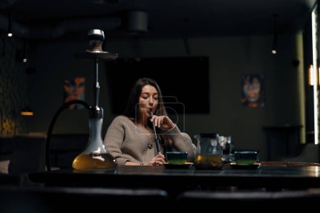 Photo for Beautiful young woman inhaling hookah girl smoking hookah sitting in lounge bar concept relaxation and smoking - Royalty Free Image
