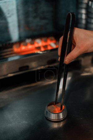Téléchargez les photos : Charbon pour narguilé est rôti sur le feu le travailleur narguilé tourne le charbon avec des pinces spéciales - en image libre de droit
