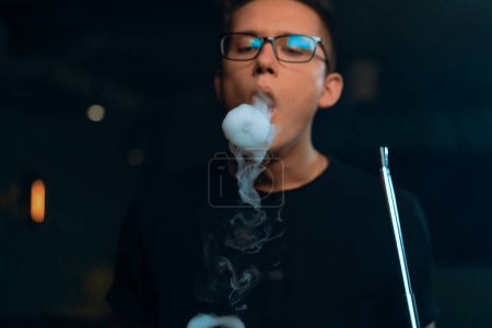 Photo for A man in glasses smokes a traditional hookah pipe A man exhales rings of thick smoke in hookah cafe or lounge bar - Royalty Free Image