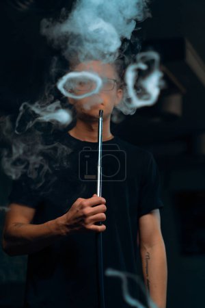 Ein Mann mit Brille raucht eine traditionelle Wasserpfeife Ein Mann atmet in einem Shisha-Café oder einer Lounge-Bar dicke Rauchschwaden aus