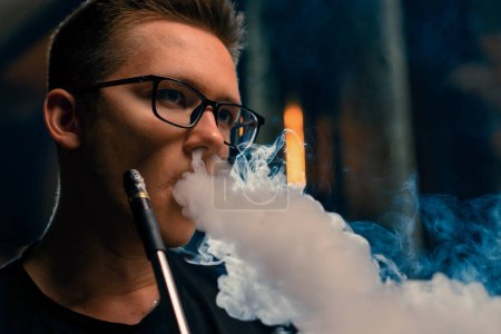 Photo for A man in glasses smokes a traditional hookah pipe A man exhales thick smoke in hookah cafe or lounge bar - Royalty Free Image