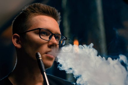 Foto de Un hombre con gafas fuma una pipa tradicional de narguile Un hombre exhala humo espeso en la cafetería de narguile o en el bar salón - Imagen libre de derechos