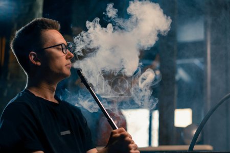 Photo for A man in glasses smokes a traditional hookah pipe A man exhales thick smoke in hookah cafe or lounge bar - Royalty Free Image