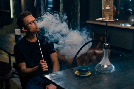 Foto de Un hombre con gafas fuma una pipa tradicional de narguile Un hombre exhala humo espeso en la cafetería de narguile o en el bar salón - Imagen libre de derechos