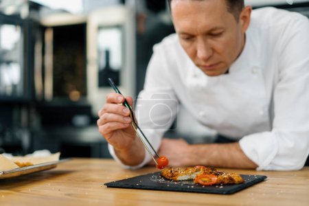 Téléchargez les photos : Chef cuisinier dans la cuisine professionnelle pose tomates cerises avec pinces morceaux frits de filet de poulet dans la chapelure - en image libre de droit