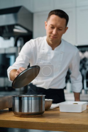 Téléchargez les photos : Cuisine professionnelle chef satisfait le chef soulève le couvercle de la casserole et vérifie l'état de préparation de la cuisson des plats - en image libre de droit