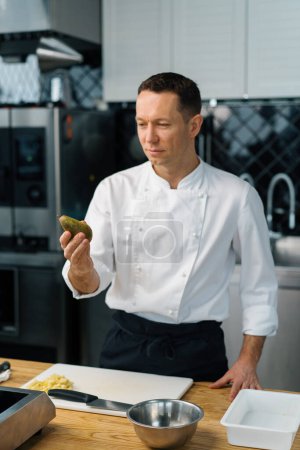 Photo for Professional kitchen satisfied chef holding pear in hand and looking cooking concept - Royalty Free Image