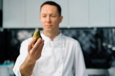 Photo for Professional kitchen satisfied chef holding pear in hand and looking cooking concept - Royalty Free Image