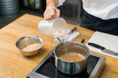 Téléchargez les photos : Cuisinier professionnel verse de l'eau dans une casserole cuisson avoine petit déjeuner concept de saine alimentation - en image libre de droit