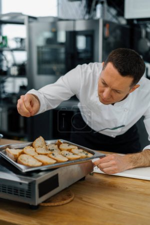 Téléchargez les photos : Un chef professionnel dans la cuisine du restaurant moderne saupoudrer d'épices sur des tranches de pain frais - en image libre de droit