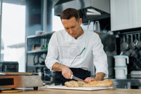 Foto de Cocina moderna profesional en restaurante chef corta pan recién horneado listo del horno tostado con cuchillo - Imagen libre de derechos
