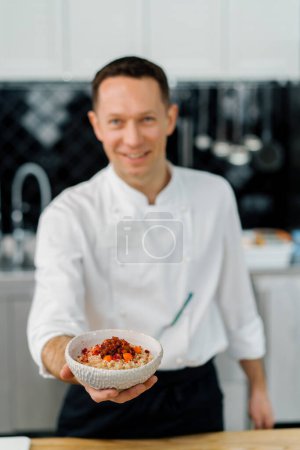 Téléchargez les photos : Cuisine professionnelle chef satisfait montrant fraîchement préparé flocons d'avoine avec petit déjeuner aux fruits caramélisés close-up - en image libre de droit