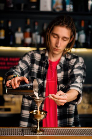 Foto de El camarero vierte el cóctel terminado en un hermoso vaso de coctelera a través de un colador de alcohol - Imagen libre de derechos