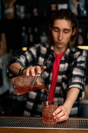 Téléchargez les photos : Barman expérimenté versant negroni cocktail alcoolisé avec des cubes de glace en cristal de mesurer le verre dans une barre de verre - en image libre de droit