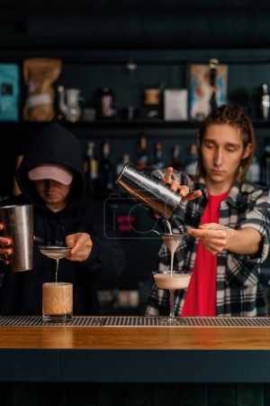 Foto de Dos camareros jóvenes preparando cócteles alcohólicos en el bar mezclando ingredientes trabajando en parejas en la fiesta - Imagen libre de derechos
