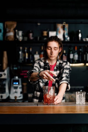 Photo for Experienced bartender stirs Negroni alcoholic cocktail with crystal ice cubes with spoon at the bar - Royalty Free Image