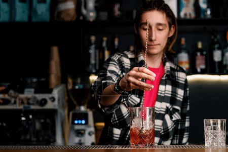 Téléchargez les photos : Un barman expérimenté remue un cocktail alcoolisé Negroni avec des glaçons en cristal avec une cuillère au bar - en image libre de droit