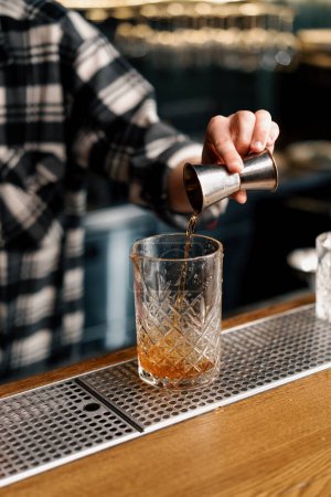Téléchargez les photos : Un barman verse de l'alcool fort d'une gigue en acier dans un verre dans le processus de fabrication de cocktail - en image libre de droit