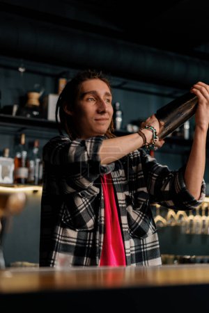 Téléchargez les photos : Un jeune barman aux dreadlocks mélange les ingrédients d'un cocktail alcoolisé en secouant le shaker - en image libre de droit