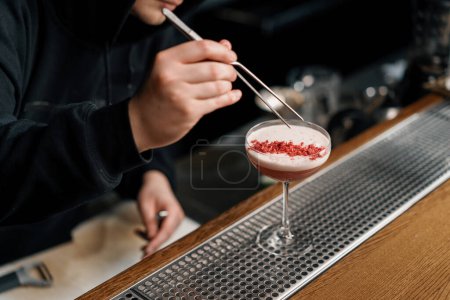 Foto de El camarero prepara un cóctel de ponche de leche y pone frambuesas picadas encima de la bebida con pinzas - Imagen libre de derechos