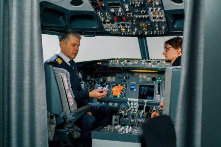 Téléchargez les photos : Un pilote expérimenté instruit un jeune élève et montre un petit avion modèle dans le poste de pilotage d'un simulateur d'avion - en image libre de droit