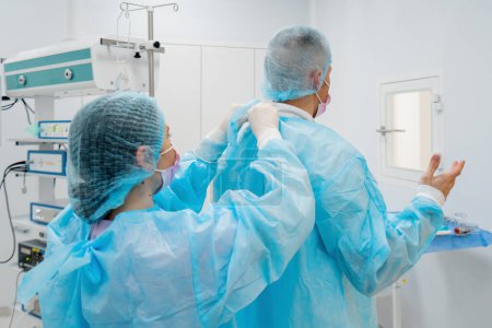 Foto de La enfermera ayuda al cirujano a ponerse una bata estéril antes de la cirugía uniforme en la preparación del quirófano para la cirugía. - Imagen libre de derechos