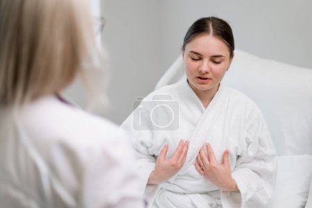 Foto de Una paciente joven se queja al médico sobre el estado de su salud acerca del dolor torácico en la sala del hospital - Imagen libre de derechos