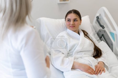 Téléchargez les photos : Une femme médecin debout près du lit dans une chambre d'hôpital et discutant d'un plan de traitement avec une jeune patiente - en image libre de droit