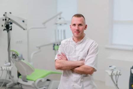Téléchargez les photos : Portrait d'un beau jeune dentiste souriant médecin debout dans le cabinet dentaire avant de commencer la procédure - en image libre de droit