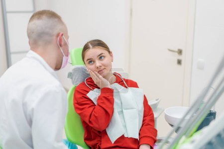 Foto de Paciente femenina molesta en la cita con el dentista se queja dolor de muelas y caries sosteniendo sus mejillas consultorio dental - Imagen libre de derechos