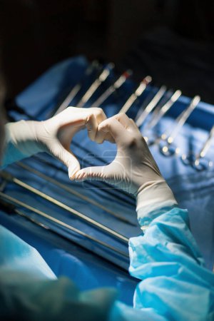 Foto de El médico muestra el signo cardíaco con las manos después de la operación en el fondo de los instrumentos quirúrgicos - Imagen libre de derechos