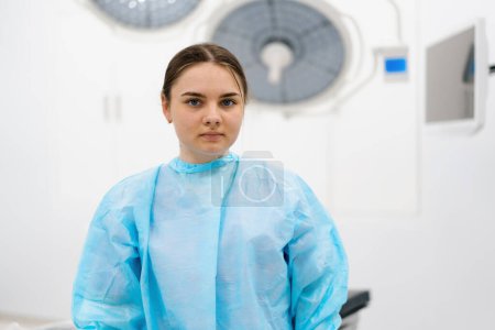 Photo for Portrait of a young female doctor in a mask and sterile protective clothing standing in the operating room after  operation - Royalty Free Image