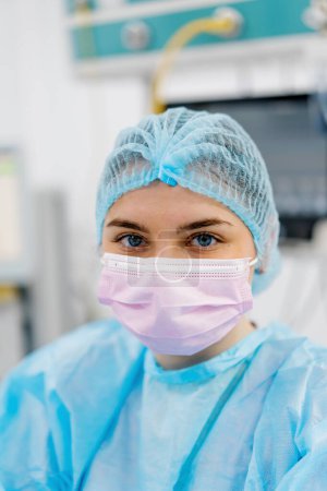 Foto de Retrato de una joven doctora con máscara y ropa protectora estéril de pie en el quirófano después de la operación - Imagen libre de derechos