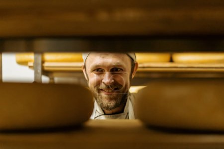Téléchargez les photos : Fromager en uniforme à l'homme de production de fromage dans l'entrepôt avec des étagères en bois têtes pleines de fromage choisit fromage - en image libre de droit