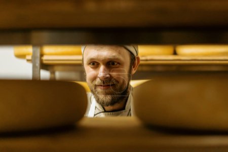 Foto de Fabricante de queso en uniforme en el hombre de producción de queso en el almacén con estantes de madera cabezas llenas de queso elige queso - Imagen libre de derechos